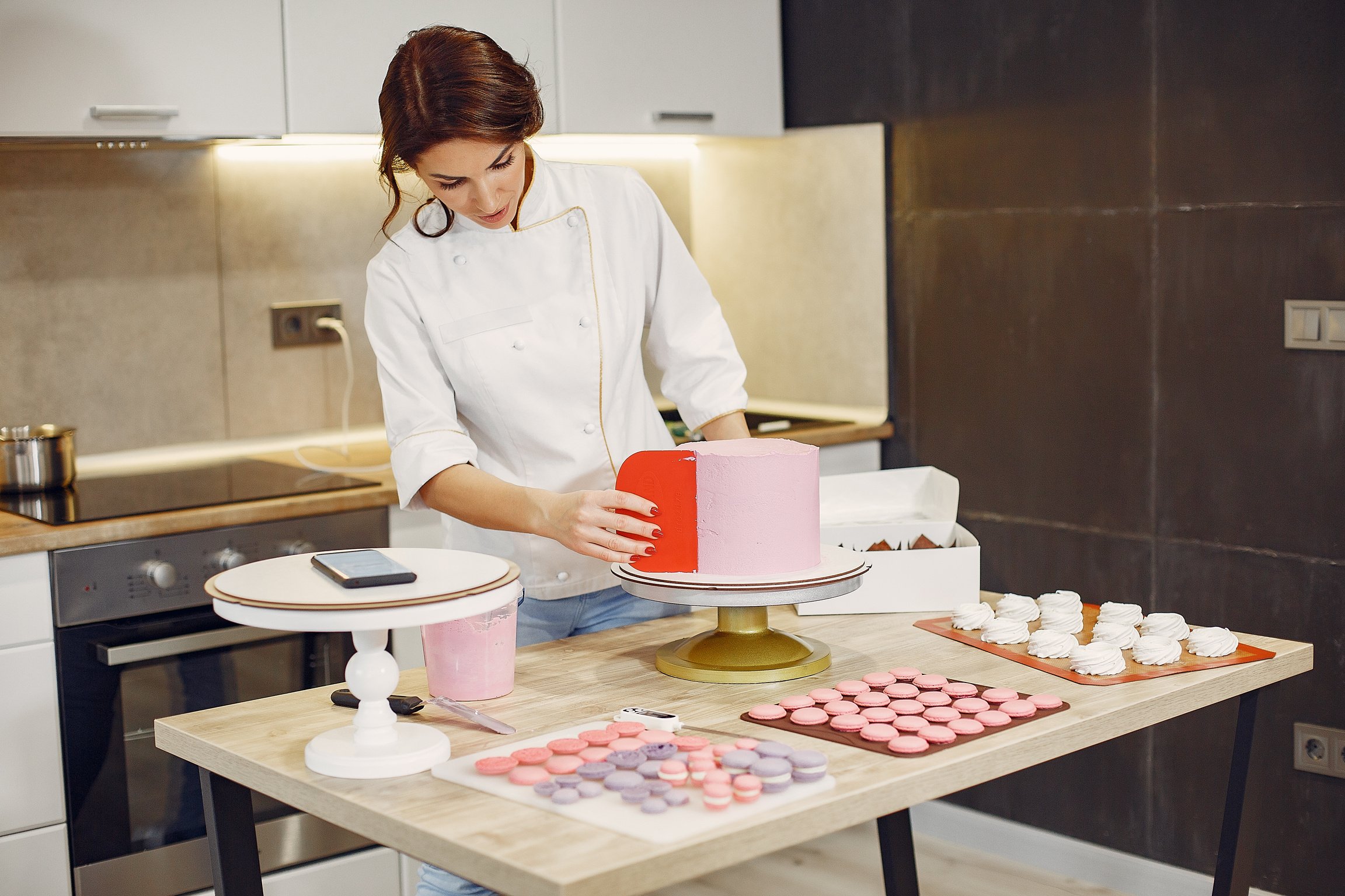 Focused female pastry chef preparing cake and desserts with smartphone aside in modern kitchen